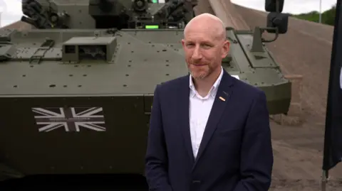 A bald man in a navy suit and white open-collared shirt smiles at the camera, he stands in front of a green armoured vehicle on a concrete surface.