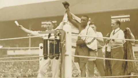 A black-and-white image of a man lifting the arm of a boxer in the ring