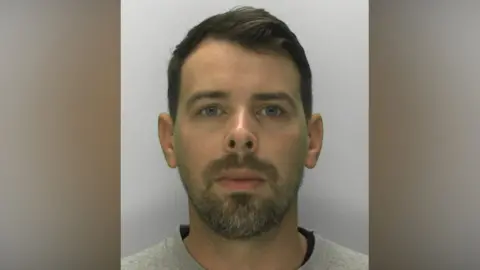 Gloucestershire Constabulary James Blandford wearing a grey T-shirt and standing against a plain grey background, looking at the camera with a blank expression.