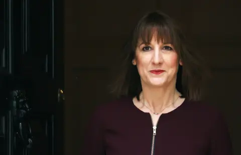 Rachel Reeves leaves a home through a black door. She is wearing a dark purple jumpsuit, with a gold necklace. She has dark hair style in a bob. 