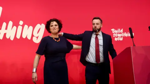 Pacemaker Claire Hanna and Colum Eastwood standing on stage side by side, both smiling. There is a red wall behind when with "build something new" written in white.