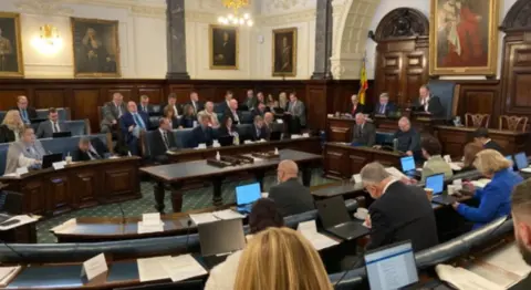 A formal looking room with wooden tables. There are benches on which people are sat looking at laptops. A man in formal robes with a chain is presiding from a larger chair he is flanked by other men in suits. 