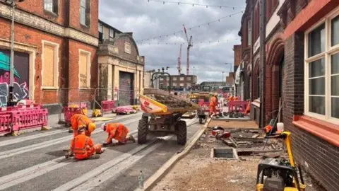 Three construction workers in orange jumpsuits work on the tram tracks on Meriden Street, with a vehicle next to them. More workers are in the background. 