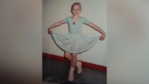 Melissa Hamilton Melissa Hamilton as a child. She is wearing a light blue leotard with light blue shirt and pink sock. The number 8 is on her leotard. Her hair is slicked back and she is holding her shirt up at the sides and her right leg is bend backwards in a curtsey position.