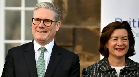 PA Media A man with grey hair and glasses wearing a suit, Sir Keir Starmer, smiles while standing next to a smiling woman with medium length brown hair and dark suit, Eluned Morgan.