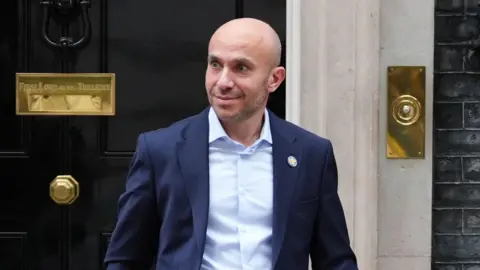 PA Media Chief Executive Officer of Morrisons Rami Baitieh leaving Downing Street. He wears a dark blue jacket over a light blue shirt with no tie and has a bald head and stubble on his chin.
