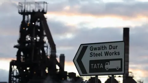 A silhouette of the steelworks machinery on a sunset sky with a road sign in the forefront which reads "steelworks, TATA".