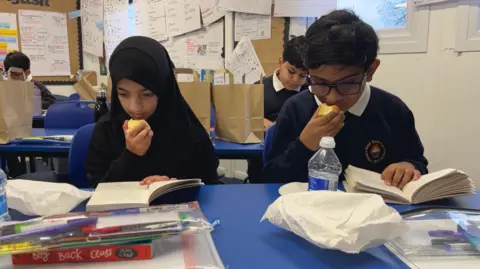 A boy and a girl sit reading while eating an apple, with pencil cases on their desk along with a water and a folder in front of them. In the background, are kids also reading and paper bags which they came in.