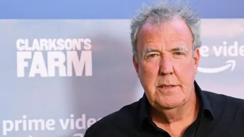 Jeremy Clarkson, a man with short grey hair wearing a black collared shirt, is pictured looking at the camera. Behind him, on a board, are the words "Clarkson's Farm" and "Prime Video". He appears to be at a promotional event. 