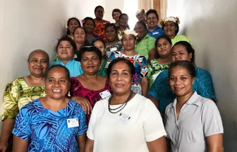 UNDP Dr Puakena Boreham, tengah, mengenakan pakaian putih, dikelilingi oleh 20 wanita, semuanya tersenyum, beberapa mengenakan pakaian tradisional Tuvalu dengan bunga di rambut mereka, dalam foto dari tahun 2018