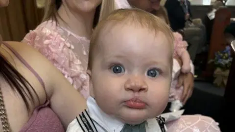 A baby, Eli, pictured in a church. He is wearing a white skirt, a blue bow tie and black suspenders. He has blonde hair and is sitting on a woman's lap, she is wearing a pink spaghetti strap dress. The woman next to them is wearing a pink dress.