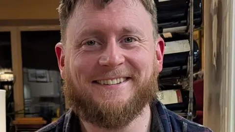 A man with a brown beard is smiling looking at the camera. He is wearing a checkered shirt.