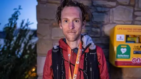 Will Roberts/willbaldlygo A man with dark hair is looking into the camera with a slight smile. He is wearing a red coat and an orange lanyard, and black rucksack straps are over his shoulders. Behind him is a yellow defibrillator box.