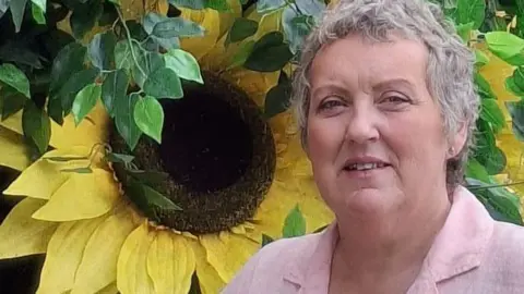PA Media Tracy Tomlinson with short grey hair wearing a pale pink cotton shirt. She is standing beside huge yellow sunflowers.