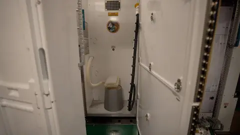 Kevin Church/BBC Toilet at Johnson Space Centre where astronauts train. The door is open to a small cubicle revealing a suction powered space station toilet. On the ground is a cylindrical grey base and on top of this is a white flat plastic oblong lid and seat. Coming out of this on the left hand side of the picture is a white corrugated pipe. On the right hand side is a black pipe with a yellow funnel at the end of it which is clipped to the wall. There is a sticker on the back wall of the cubicle that says international space station orbital outhouse team with a cartoon spaceman next to an old fashioned toilet. 