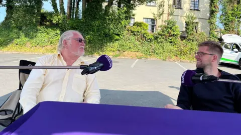 Two men arguing behind a branded purple BBC Radio Guernsey microphone. On the left is Lord Digby Jones, he has white shoulder length hair and a yellow shirt and on the right is Deputy Andy Taylor, he's wearing a blue Guernsey and glasses and has spiky blonde hair and a beard.