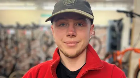 A young man in a green baseball cap, black T-shirt and red fleece jacket is smiling into the camera. There are bikes in the background.