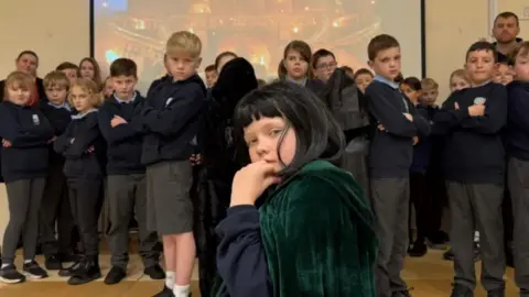 A group of school children with their arms folded with one boy in the front in a black wig