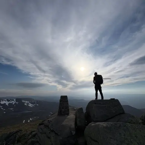 Rory Grant A man silhouetted at the summit of a climb, with hills and sky stretching out in front of him.