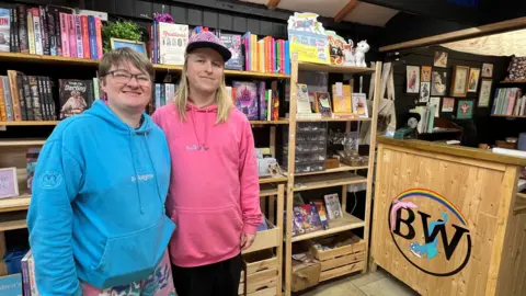 Miles Nelson and Chis Lameth smile in front of stacked book shelves at ther stall. Miles has short light brown hair and wears a blue hoody and glasses. Chris has long blonde hair and wears a pink hoody and a cap. Both hoodies have Bookwyrm's logo on them.