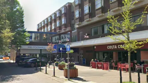 Google Exterior view of the Britannia Ashley Hotel in Hale in Trafford. A small blue marquee entrance to the building is visible, next to a Costa coffee shop and an M&S food hall. 