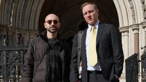 PA Media Two men stand in the centre of the image looking at the camera. Carlo stands on the left, wearing sunglasses and a long puffer coat, while Tom stands on the right wearing a charcoal grey suit with a pale yellow tie.