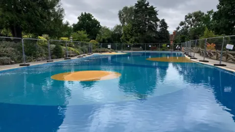 A public paddling pool with blue and yellow flooring. It is surrounded by a footpath and trees