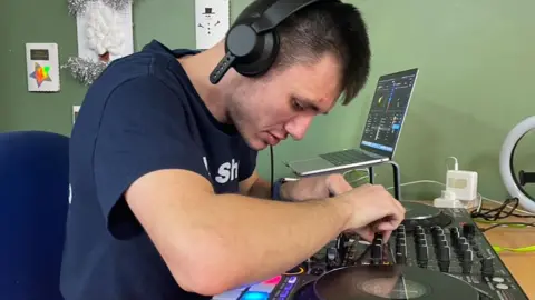 Jacob Towner is visually impaired. He is wearing a headset and sitting at a table operating a mixing desk to prepare his music for his Saturday night radio show.