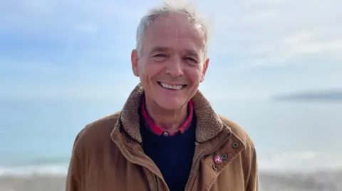 Richard Henthorn is an older gentleman, with short grey hair. He is smiling and wearing a brown over a jumper and shirt. He is standing in front of a shoreline on a sunny day.
