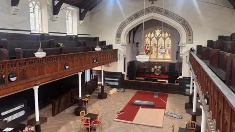 The inside of the church from the balcony, which runs around three of the four walls. There is an archway at the far wall with the stage on it and a stained glass window. Below is a wooden floor with seating and small tables. On the balcony are rows of wooden pews