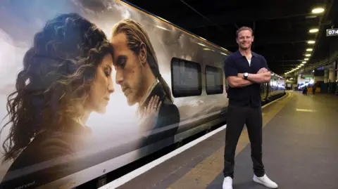 PA Media Sam Heughan stands in front of a train. He has a navy blue shirt and he is crossing his arms. The train has a picture from TV show Outlander on it, comprised of a woman with long brown curly hair embracing a man with long blonde hair.