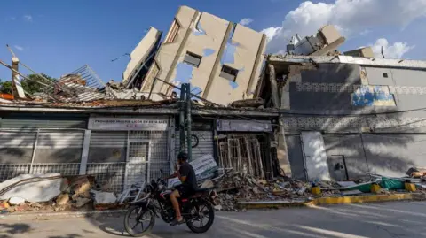 MIGUEL GUTIERREZ/EPA/Shutterstock A person on a motorbike looks at buildings destroyed by earthquakes in Catia La Mar on 14 July 2026. Part of a building lies toppled on the remains of another. Rubble is piling up in the street. 