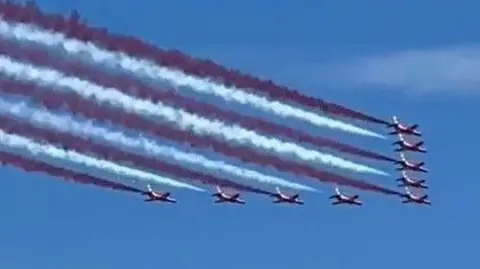 Red arrows in formation with red, white and blue smoke behind them against a blue sky