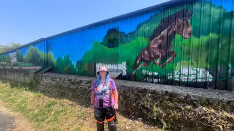 A woman smiling widely in front of a mural with a picture of a horse on it. The woman is in black trousers and a purple T-shirt with white writing on it. She is also wearing a white and pink bucket hat. The horse in the mural is jumping through the air.
