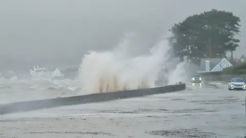 JohnRoss MacMathúna Large waves crash over a sea wall and onto a coastal road. Tow cars are driving on the road