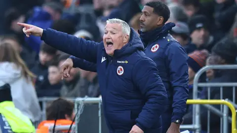Football coach Rob Kelly standing on a sideline in a stadium pointing with his outstretched right hand while wearing a dark blue coat with the Reading badge on its chest, another coaching staff member stands behind with the crowd in the background