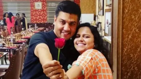 Sunil and Priya Rastogi are hugging in a restaurant and smiling at the camera. Sunil is holding up a rose to the camera.