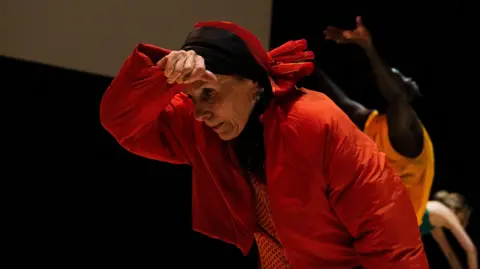 Luca Truffarelli Joanna Banks. She is wearing orange clothing and a black and orange headscarf. She is bent over slightly, holding her right arm up to her forehead. Other dancers are behind her.