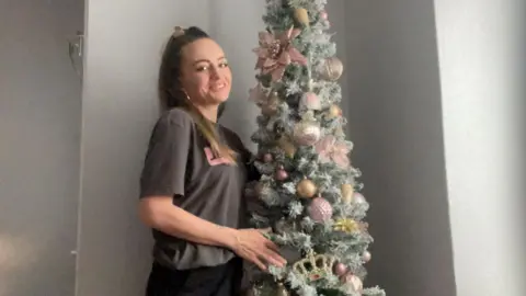 Sally-Anne Long, a woman with her hair tied back wearing a grey t-shirt, stands next to a Christmas tree decorated with gold and red ornaments.