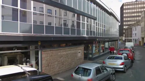 A picture of Dumaresq Street. There are a number of cars parked to the right of the image. There are two lorries parked in the distance.