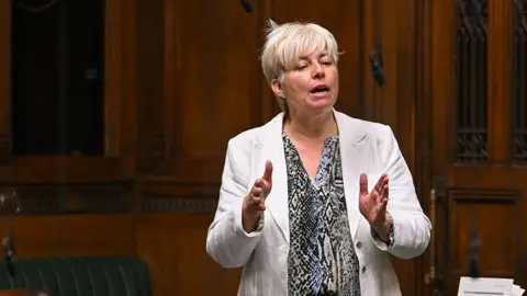 UK Parliament Lia Nici speaking in the House of Commons. She has short blond hair and is wearing a patterned shirt and a white jacket