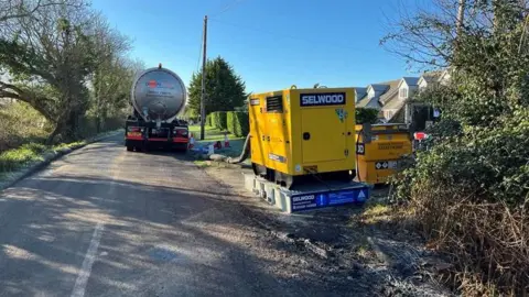 A big tanker on a country lane which has a metal tank - there is a pipe connected to a yellow generator which is connected to the pumping station. The tanker is making sure people can use their toilets and sinks 