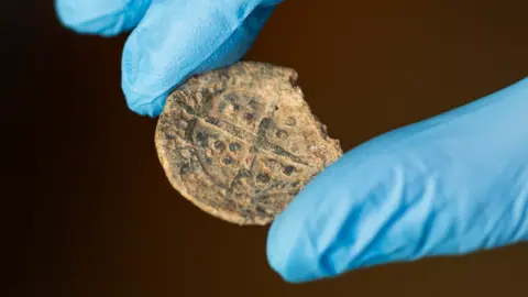 National Trust A close-up of a medieval St Nicholas or boy bishop token, held between the thumb and forefinger of a hand wearing blue gloves. The token is damaged on the right side. On its front is a cross with three small dots within each angle of the cross. 