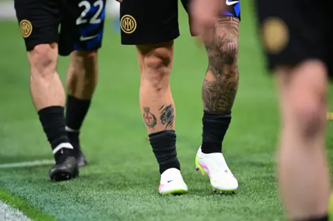 A close up of a footballer's legs he has tattoos on both calves. He is wearing black socks, white trainers and black shorts.