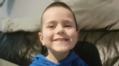 Family handout Kyran Durnin - a young boy with short, brown hair is smiling at the camera as he sits on a dark, leather sofa. He is wearing a blue hoodie.