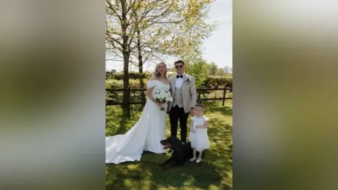 Kennedy News Ashleigh Stanley and Jake Stanley. She stand on the left in a wedding dress. He is on the right in black trousers, a beige jacket and waistcoat. Next to him is a small girl and a dobermann 