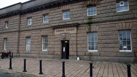 A general view of a sandstone building with a paved area in front of a doorway, over which is a sign reading 'Crown Court'.