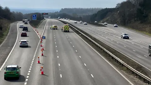 BBC M1 coned off. Vehicles are going down a slip road while a police vehicle and ambulance are in one lane of the motorway. The other carriageway has a number of vehicles on. 