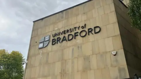 A large building carrying a sign reading: "The University of Bradford." The wall also has a university symbol, split into four sections. In the background there is a tree and a security camera. 