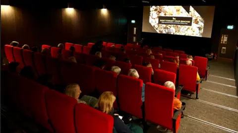 The Maltings Trust Rows of red seats make up a small cinema seating about 70 people. Most of the seats are taken and an advert is on the screen. 